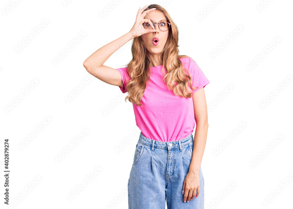 Young beautiful caucasian woman with blond hair wearing casual clothes and glasses doing ok gesture shocked with surprised face, eye looking through fingers. unbelieving expression.