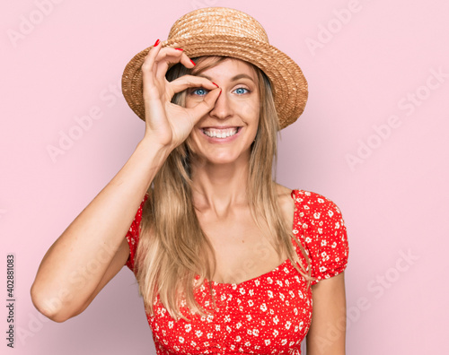 Beautiful caucasian woman wearing summer hat doing ok gesture with hand smiling, eye looking through fingers with happy face.