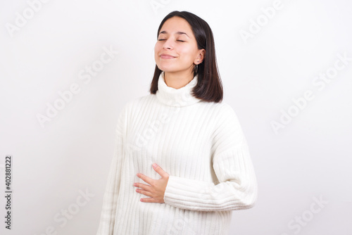 Fotografie Young brunette woman wearing white knitted sweater against white background touches tummy, smiles gently, eating and satisfaction concept