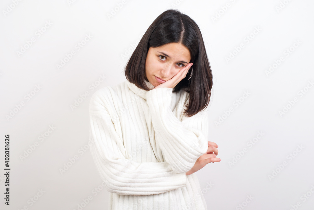 Very bored Young brunette woman wearing white knitted sweater against white background holding hand on cheek while support it with another crossed hand, looking tired and sick,