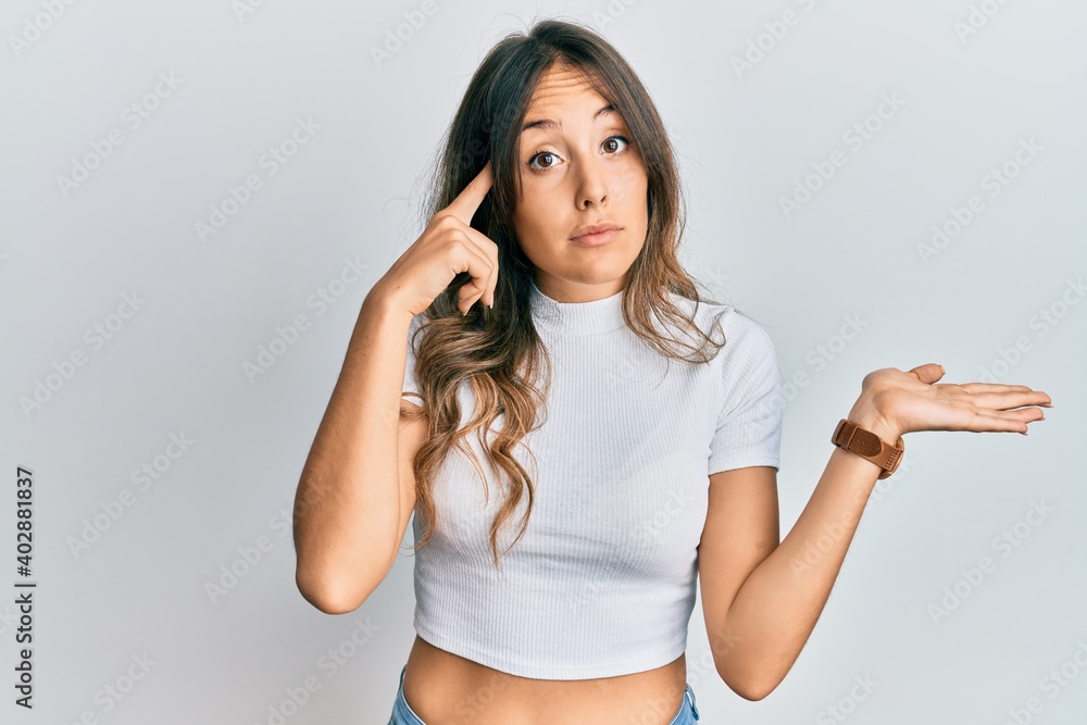 Young brunette woman wearing casual white t shirt confused and annoyed with open palm showing copy space and pointing finger to forehead. think about it. Young brunette woman wearing casual white t shirt confused and annoyed with open palm showing copy space and pointing finger to forehead. think about it.