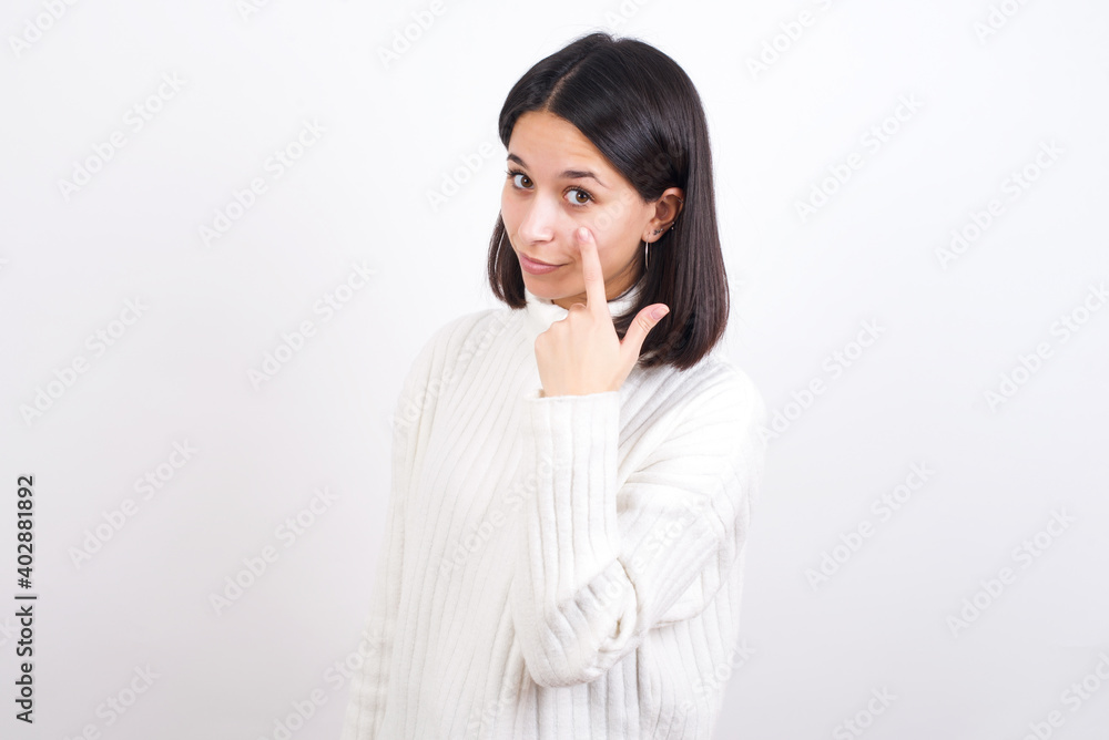 Young brunette woman wearing white knitted sweater against white background Pointing to the eye watching you gesture, suspicious expression. Young brunette woman wearing white knitted sweater against white background Pointing to the eye watching you gesture, suspicious expression.