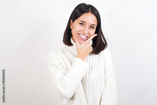 Young brunette woman wearing white knitted sweater against white background looking confident at the camera smiling with crossed arms and hand raised on chin. Thinking positive.