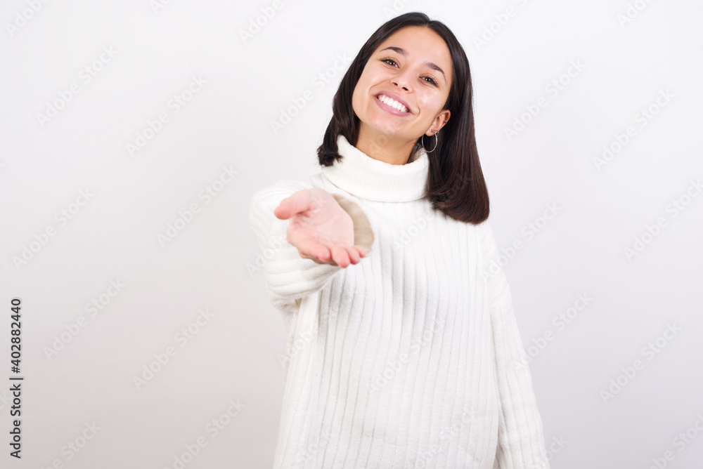 Young brunette woman wearing white knitted sweater against white background smiling friendly offering handshake as greeting and welcoming. Successful business. Young brunette woman wearing white knitted sweater against white background smiling friendly offering handshake as greeting and welcoming. Successful business.