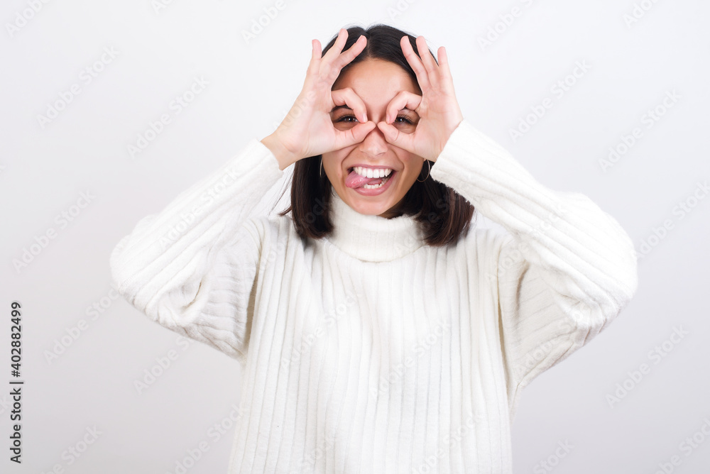 Young brunette woman wearing white knitted sweater against white background doing ok gesture like binoculars sticking tongue out, eyes looking through fingers. Crazy expression. Young brunette woman wearing white knitted sweater against white background doing ok gesture like binoculars sticking tongue out, eyes looking through fingers. Crazy expression.