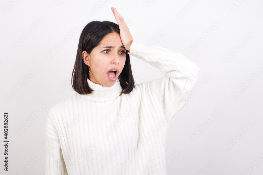 Young brunette woman wearing white knitted sweater against white background surprised with hand on head for mistake, remember error. Forgot, bad memory concept. Young brunette woman wearing white knitted sweater against white background surprised with hand on head for mistake, remember error. Forgot, bad memory concept.