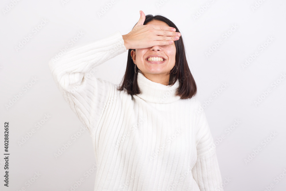 Young brunette woman wearing white knitted sweater against white background smiling and laughing with hand on face covering eyes for surprise. Blind concept. Young brunette woman wearing white knitted sweater against white background smiling and laughing with hand on face covering eyes for surprise. Blind concept.