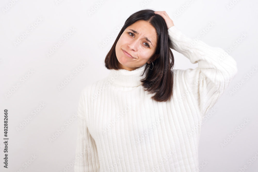 Young brunette woman wearing white knitted sweater against white background being confused and wonders about something. Holding hand on her head, uncertain with doubt. Pensive concept. Young brunette woman wearing white knitted sweater against white background being confused and wonders about something. Holding hand on her head, uncertain with doubt. Pensive concept.