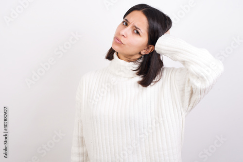 Young brunette woman wearing white knitted sweater against white background confuse and wonder about question. Uncertain with doubt, thinking with hand on head. Pensive concept. © Roquillo