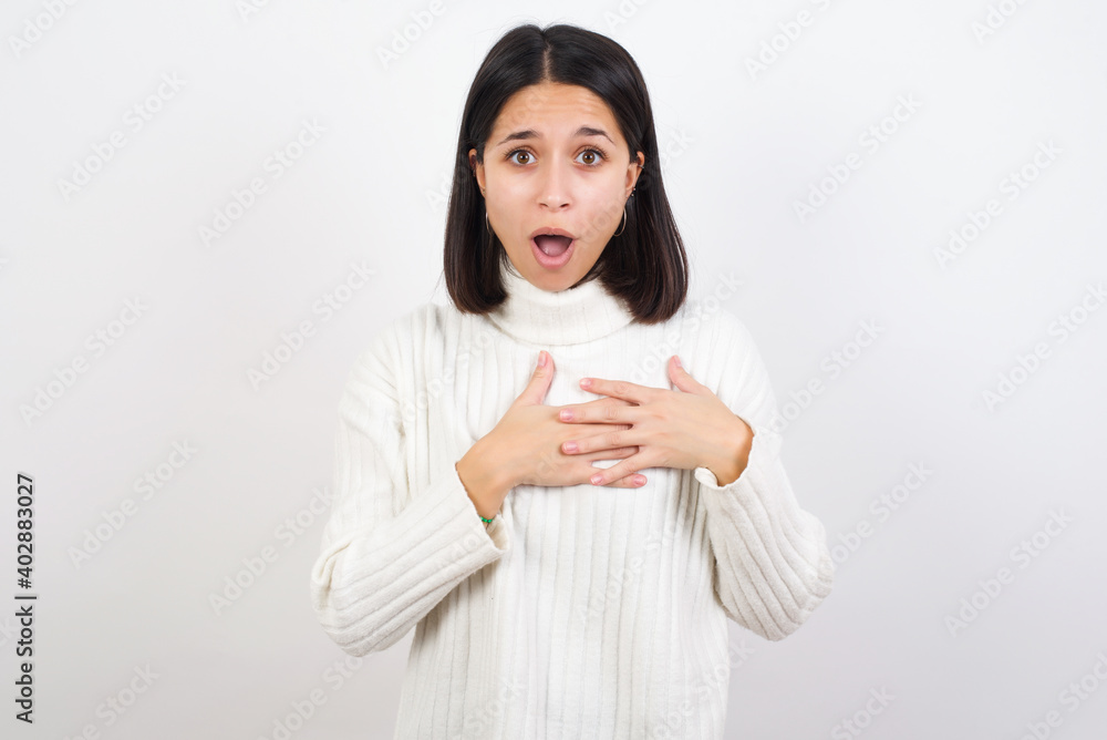 Scared Young brunette woman wearing white knitted sweater against looks with frightened expression, keeps hands on chest, being puzzled to notice something strange, People, hush reaction and emotions. Scared Young brunette woman wearing white knitted sweater against looks with frightened expression, keeps hands on chest, being puzzled to notice something strange, People, hush reaction and emotions.