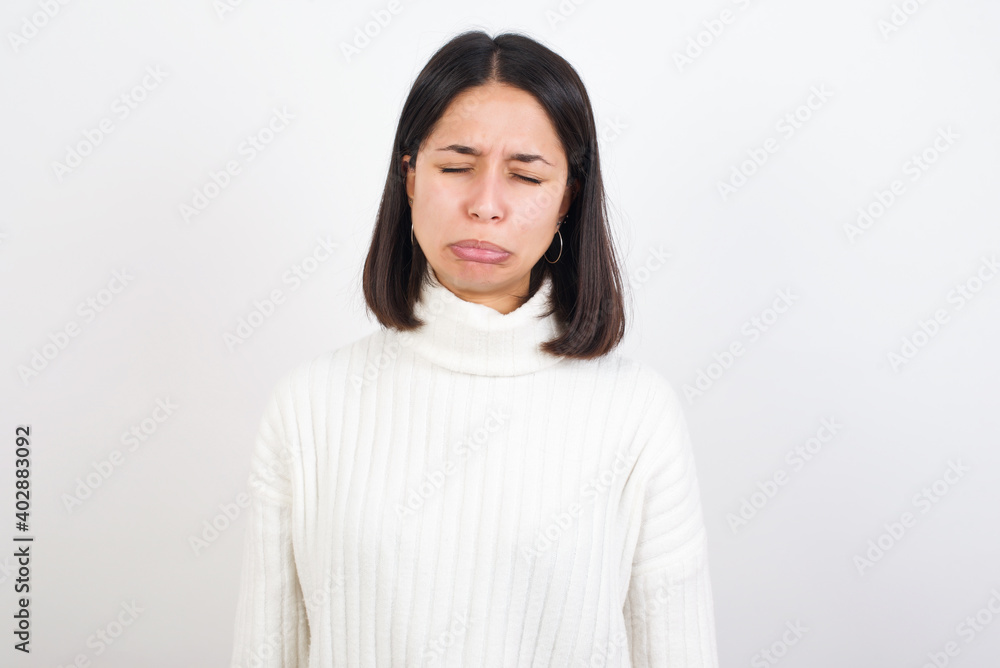 Dismal gloomy rejected Young brunette woman wearing white knitted sweater against white background has problems and difficulties, curves lower lip and closes eyes in despair, being in depression Dismal gloomy rejected Young brunette woman wearing white knitted sweater against white background has problems and difficulties, curves lower lip and closes eyes in despair, being in depression
