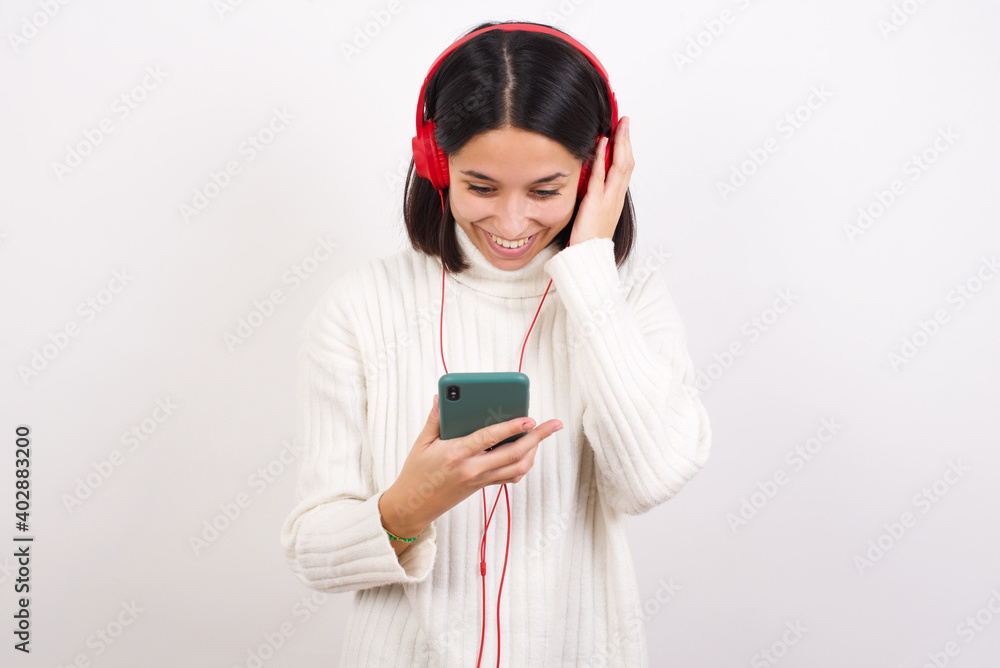 Happy Young brunette woman wearing white knitted sweater against white background feels good while focused in screen of smartphone. People, technology, lifestyle Happy Young brunette woman wearing white knitted sweater against white background feels good while focused in screen of smartphone. People, technology, lifestyle
