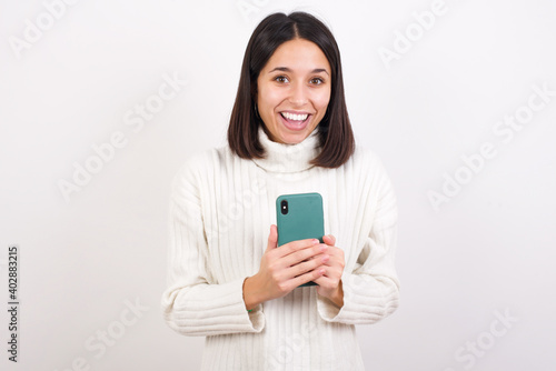 Excited Young brunette woman wearing white knitted sweater against white background holding smartphone and looking amazed to the camera after receiving good news.