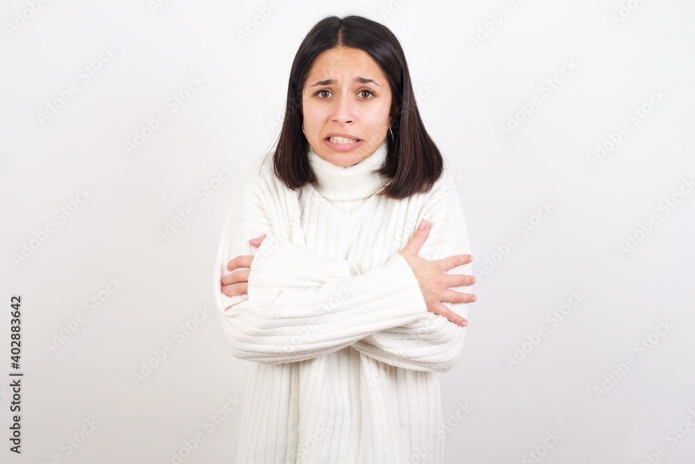 Desperate Young brunette woman wearing white knitted sweater against white background trembles and feels cold, hugs oneself to warm up or feels scared notices something terrifying. Desperate Young brunette woman wearing white knitted sweater against white background trembles and feels cold, hugs oneself to warm up or feels scared notices something terrifying.