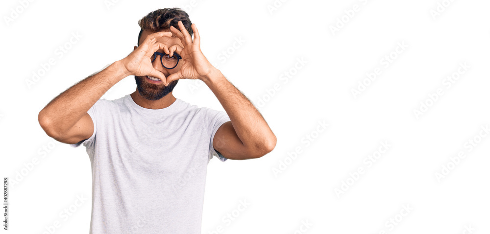 Young hispanic man wearing casual clothes and glasses doing heart shape with hand and fingers smiling looking through sign Young hispanic man wearing casual clothes and glasses doing heart shape with hand and fingers smiling looking through sign