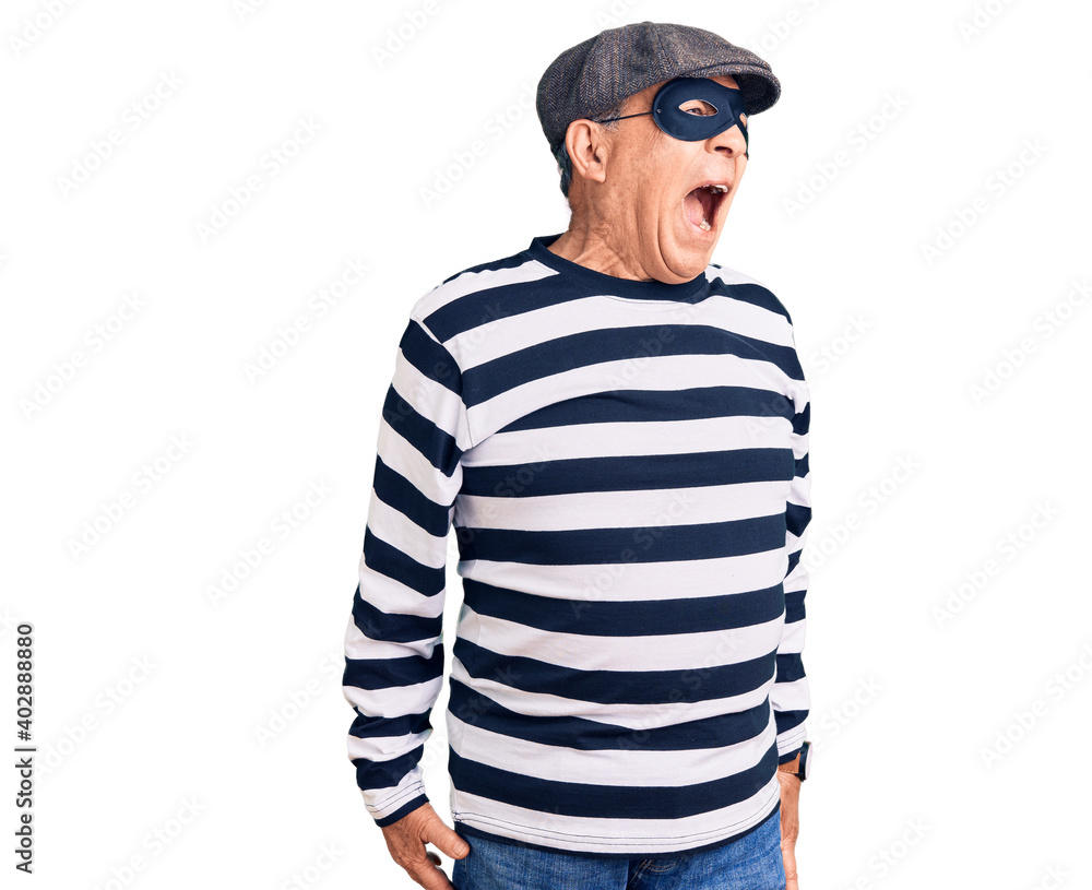 Senior handsome man wearing burglar mask and t-shirt angry and mad screaming frustrated and furious, shouting with anger. rage and aggressive concept. Senior handsome man wearing burglar mask and t-shirt angry and mad screaming frustrated and furious, shouting with anger. rage and aggressive concept.