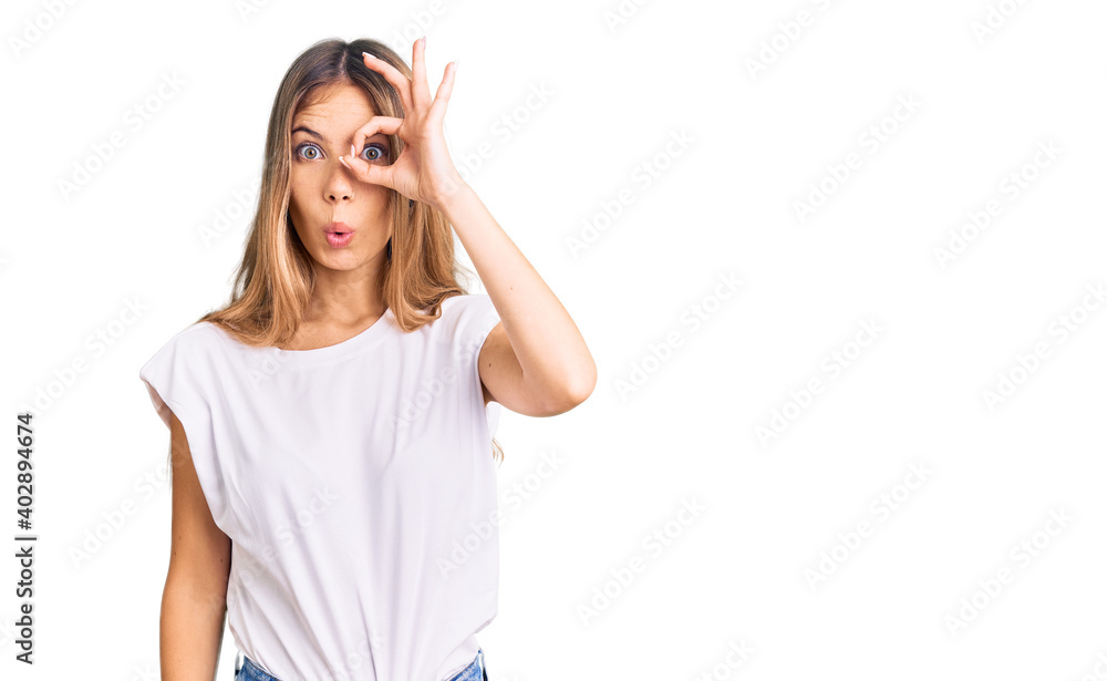 Beautiful caucasian woman with blonde hair wearing casual white tshirt doing ok gesture shocked with surprised face, eye looking through fingers. unbelieving expression. Beautiful caucasian woman with blonde hair wearing casual white tshirt doing ok gesture shocked with surprised face, eye looking through fingers. unbelieving expression.