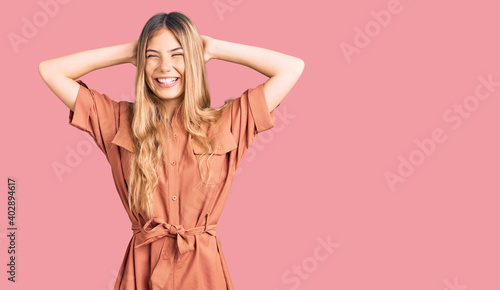 Beautiful caucasian woman with blonde hair wearing summer jumpsuit relaxing and stretching, arms and hands behind head and neck smiling happy