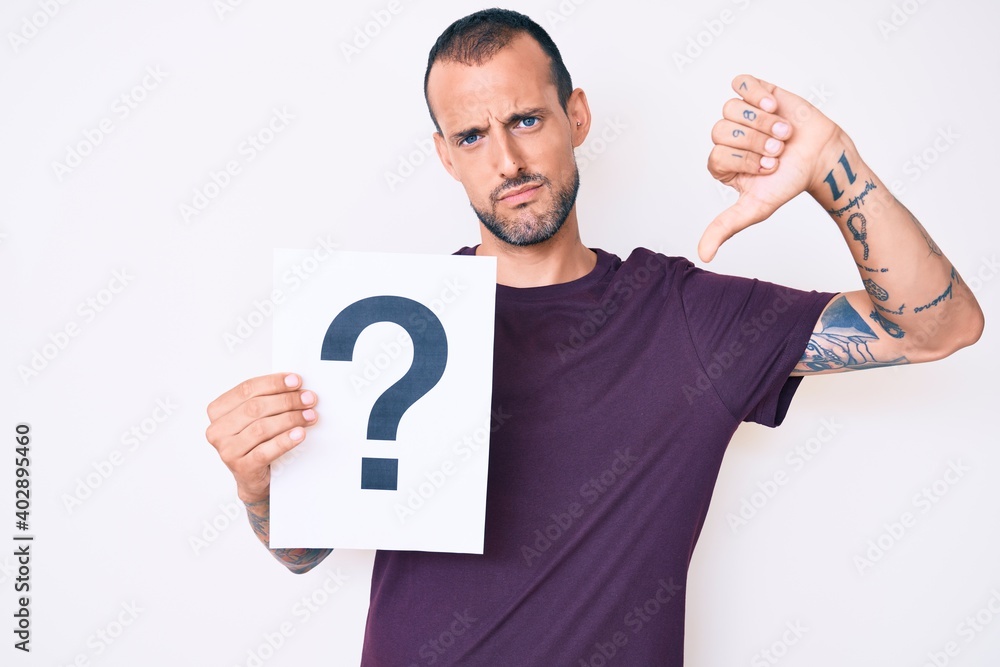 Young handsome man with tattoo holding question mark with angry face, negative sign showing dislike with thumbs down, rejection concept Young handsome man with tattoo holding question mark with angry face, negative sign showing dislike with thumbs down, rejection concept
