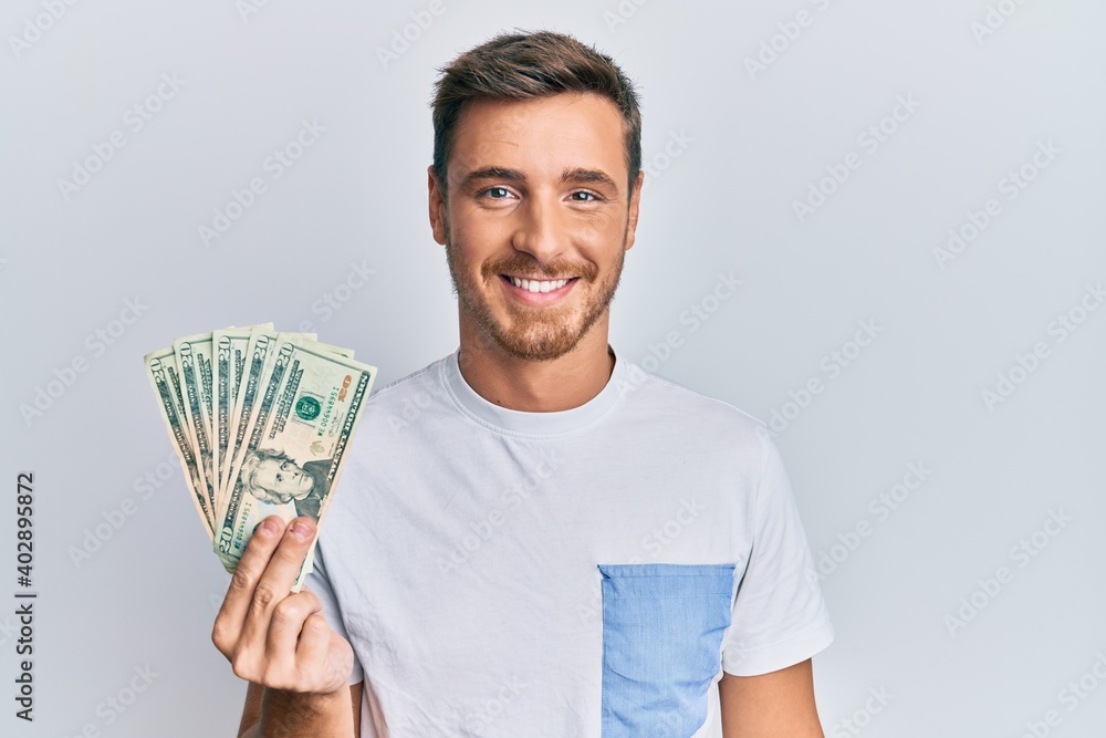 Handsome caucasian man holding 20 dollars banknote looking positive and happy standing and smiling with a confident smile showing teeth Handsome caucasian man holding 20 dollars banknote looking positive and happy standing and smiling with a confident smile showing teeth