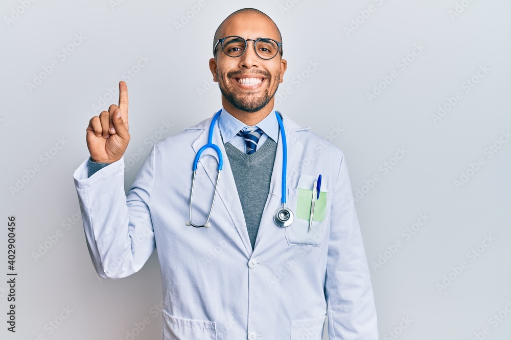 Hispanic adult man wearing doctor uniform and stethoscope showing and pointing up with finger number one while smiling confident and happy.