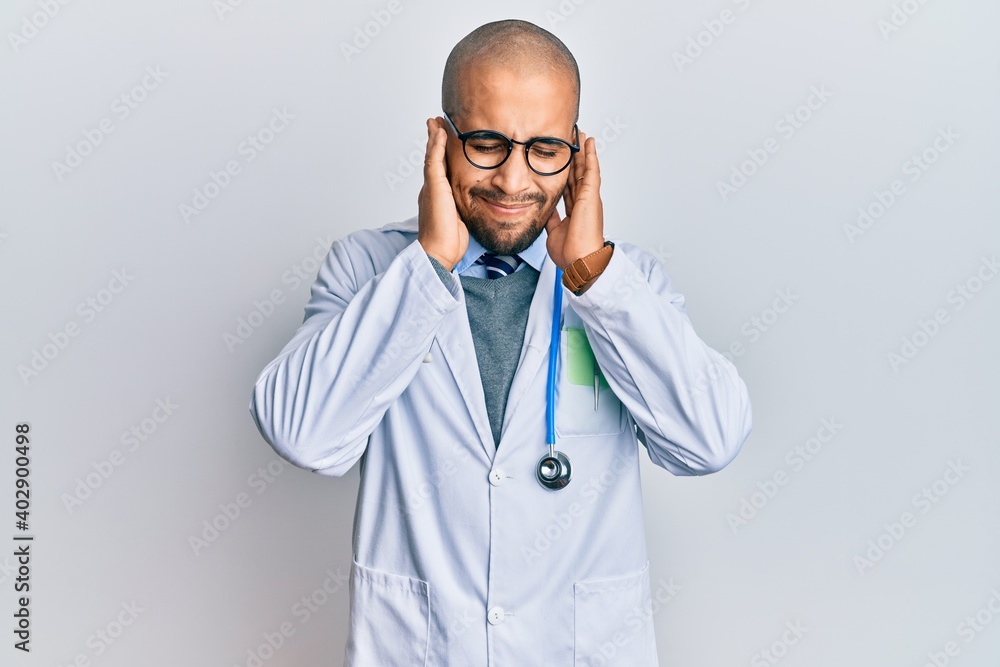 Hispanic adult man wearing doctor uniform and stethoscope covering ears with fingers with annoyed expression for the noise of loud music. deaf concept. Hispanic adult man wearing doctor uniform and stethoscope covering ears with fingers with annoyed expression for the noise of loud music. deaf concept.