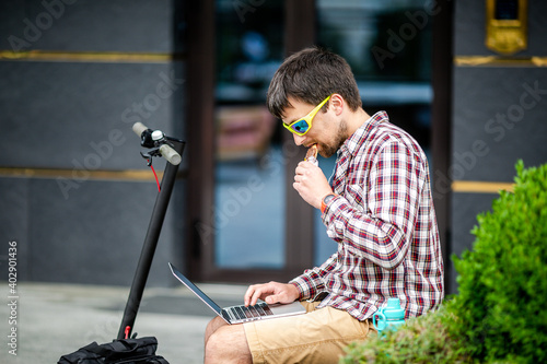 Young caucasian man working on laptop while eating nutritious bar, sitting on bench near electric scooter in yard. Healthy, fastfood. Granola bar. Muesli Bar. Protein Bar. Working from outside office photo