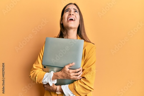 Young beautiful woman holding laptop angry and mad screaming frustrated and furious, shouting with anger looking up.