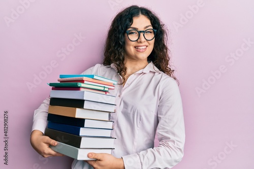 Young brunette woman with curly hair holding a pile of books smiling with a happy and cool smile on face. showing teeth.