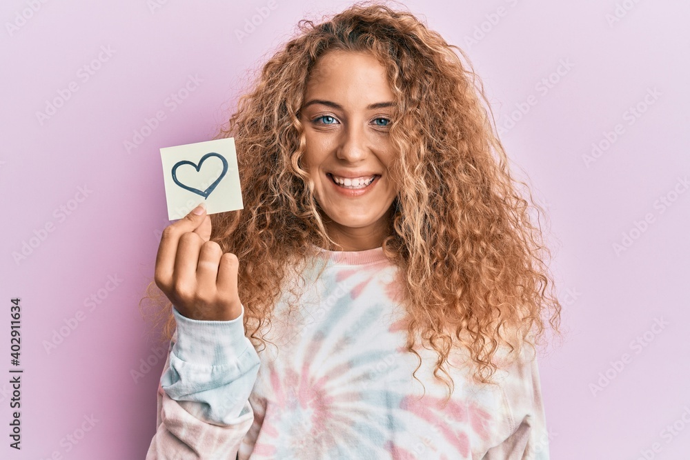 Beautiful caucasian teenager girl holding heart reminder looking positive and happy standing and smiling with a confident smile showing teeth Beautiful caucasian teenager girl holding heart reminder looking positive and happy standing and smiling with a confident smile showing teeth