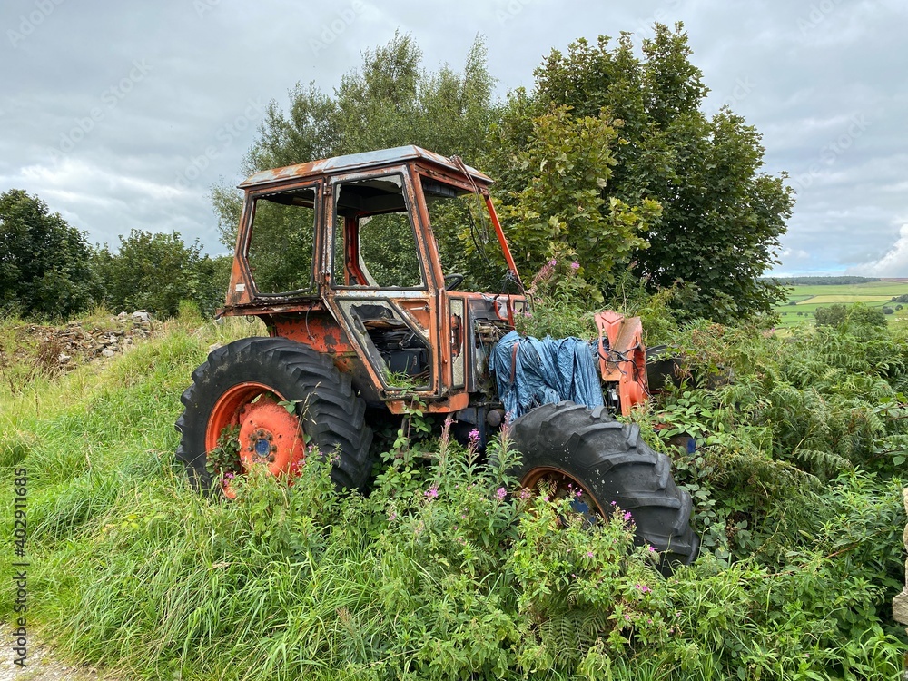 Obraz premium An old discarded orange tractor, overgrown with long grasses, with hills and meadows, in the distance near, Silsden, Keighley, UK