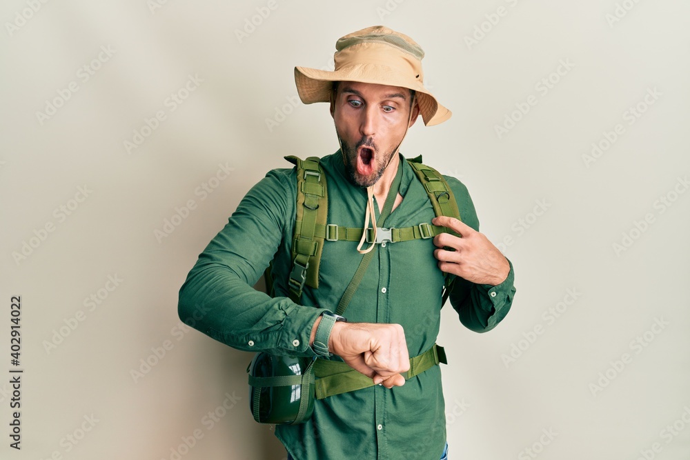 Handsome man with beard wearing explorer hat and backpack looking at the watch time worried, afraid of getting late Handsome man with beard wearing explorer hat and backpack looking at the watch time worried, afraid of getting late