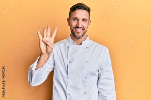 Handsome man with beard wearing professional cook uniform showing and pointing up with fingers number four while smiling confident and happy. © Krakenimages.com