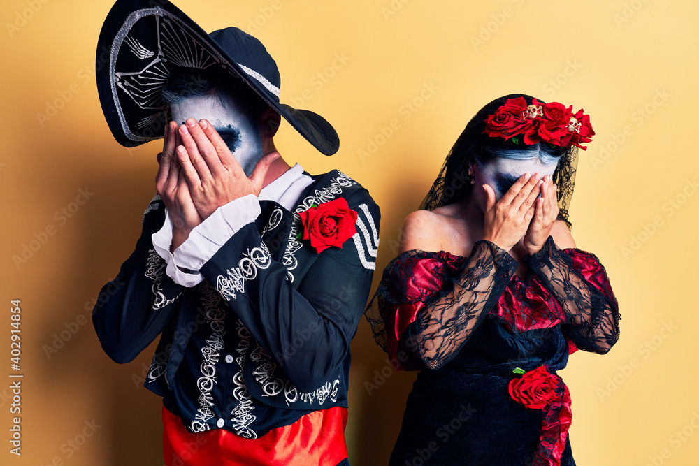 Young couple wearing mexican day of the dead costume over yellow with sad expression covering face with hands while crying. depression concept.
