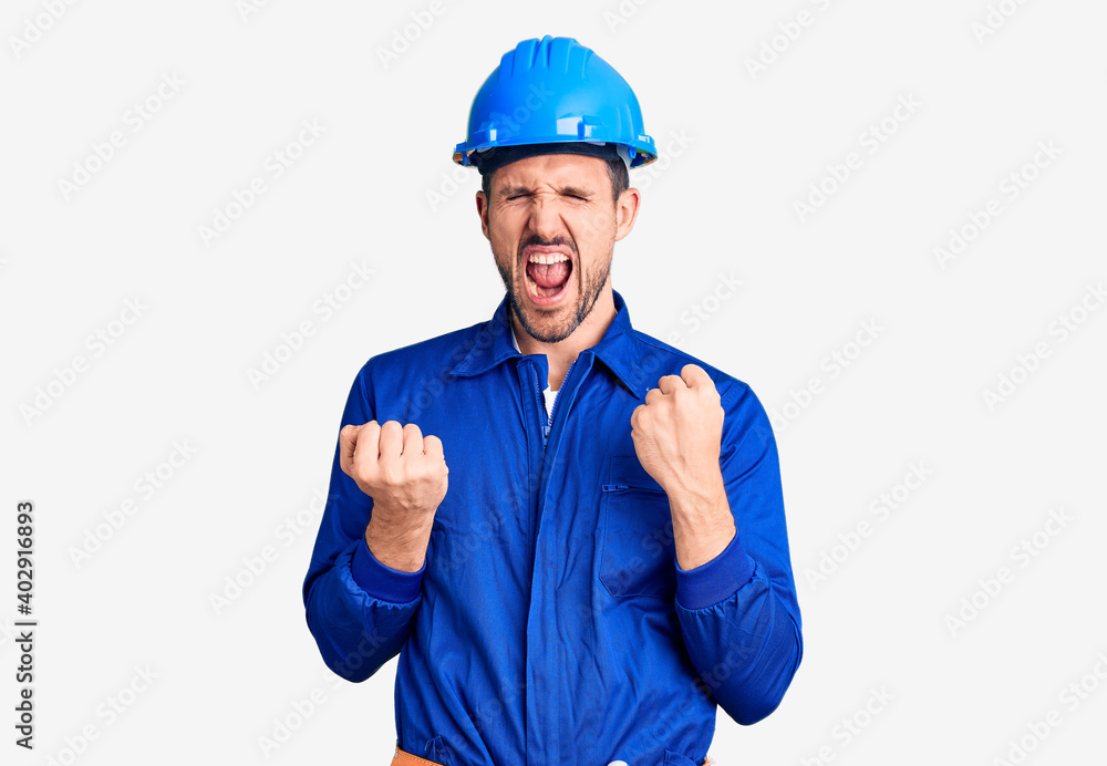 Young handsome man wearing worker uniform and hardhat celebrating surprised and amazed for success with arms raised and eyes closed. winner concept.