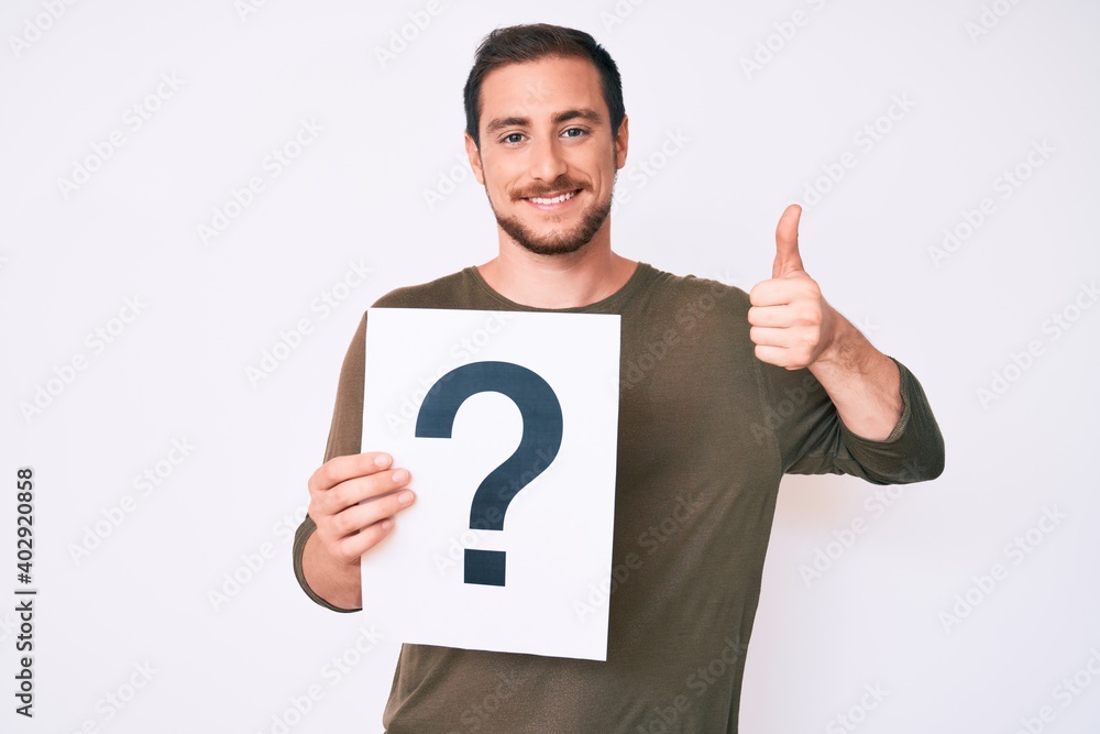 Young handsome man holding question mark smiling happy and positive, thumb up doing excellent and approval sign Young handsome man holding question mark smiling happy and positive, thumb up doing excellent and approval sign