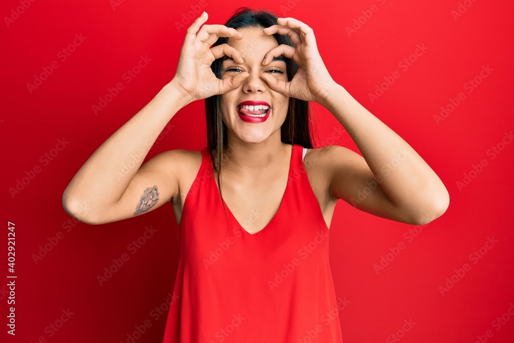 Young hispanic girl wearing casual style with sleeveless shirt doing ok gesture like binoculars sticking tongue out, eyes looking through fingers. crazy expression. Young hispanic girl wearing casual style with sleeveless shirt doing ok gesture like binoculars sticking tongue out, eyes looking through fingers. crazy expression.