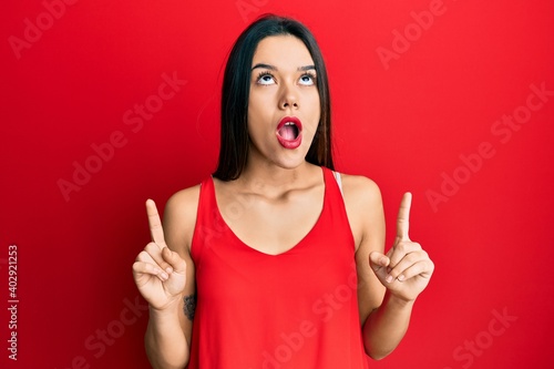 Young hispanic girl wearing casual style with sleeveless shirt amazed and surprised looking up and pointing with fingers and raised arms.