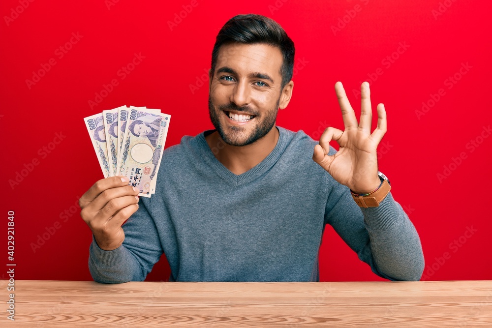 Handsome hispanic man holding japanese yen banknotes doing ok sign with fingers, smiling friendly gesturing excellent symbol Handsome hispanic man holding japanese yen banknotes doing ok sign with fingers, smiling friendly gesturing excellent symbol