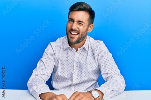 Handsome hispanic man wearing business clothes sitting on the table winking looking at the camera with sexy expression, cheerful and happy face.