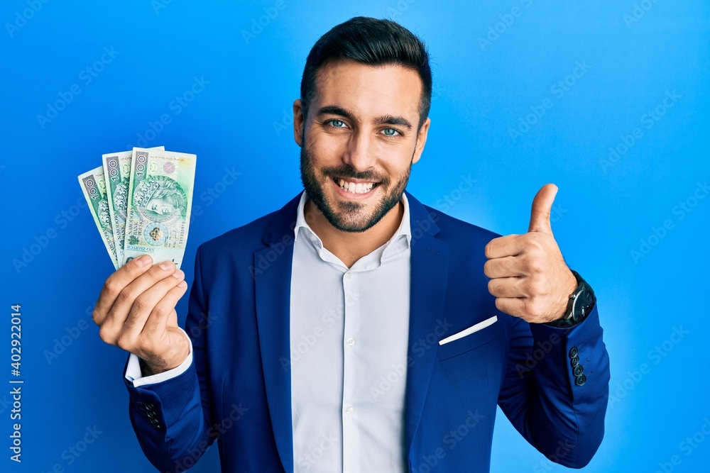 Young hispanic businessman wearing business suit holding polish zloty banknotes smiling happy and positive, thumb up doing excellent and approval sign Young hispanic businessman wearing business suit holding polish zloty banknotes smiling happy and positive, thumb up doing excellent and approval sign