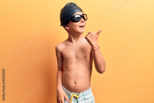 Cute blond kid wearing swimwear and swimmer glasses pointing thumb up to the side smiling happy with open mouth © Krakenimages.com