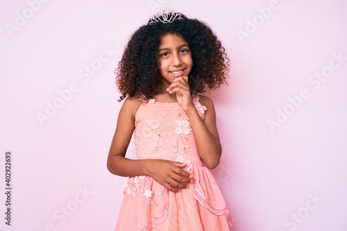 African american child with curly hair wearing princess crown smiling looking confident at the camera with crossed arms and hand on chin. thinking positive.