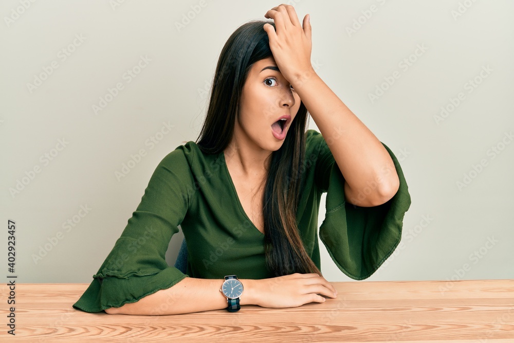 Young brunette woman wearing casual clothes sitting on the table surprised with hand on head for mistake, remember error. forgot, bad memory concept. Young brunette woman wearing casual clothes sitting on the table surprised with hand on head for mistake, remember error. forgot, bad memory concept.