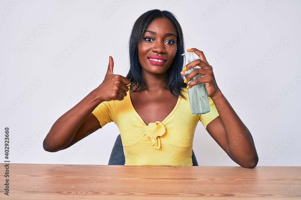 Young african american woman sitting on the table cleaning hand using sanitizer gel smiling happy and positive, thumb up doing excellent and approval sign Young african american woman sitting on the table cleaning hand using sanitizer gel smiling happy and positive, thumb up doing excellent and approval sign