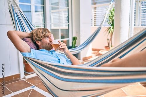 Middle age handsome man at the terrace of his house relaxing lying on a hammock drinking a fresh beer