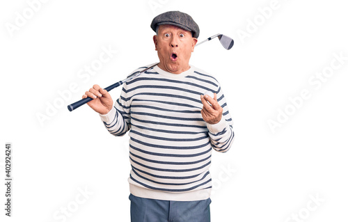 Senior handsome grey-haired man holding golf club and ball scared and amazed with open mouth for surprise, disbelief face