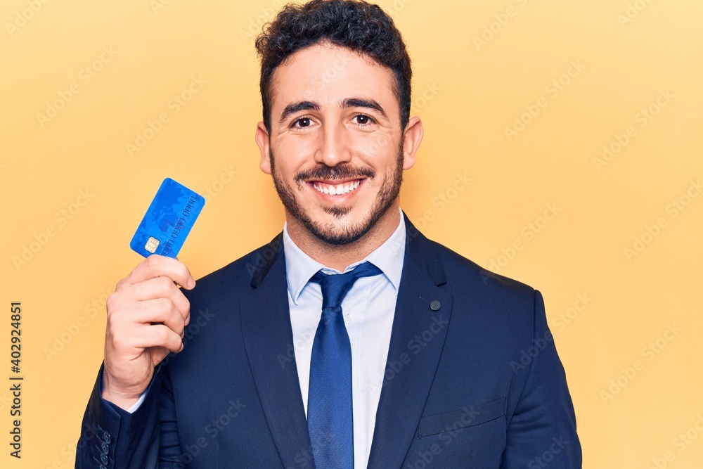 Young hispanic man wearing suit holding credit card looking positive and happy standing and smiling with a confident smile showing teeth Young hispanic man wearing suit holding credit card looking positive and happy standing and smiling with a confident smile showing teeth