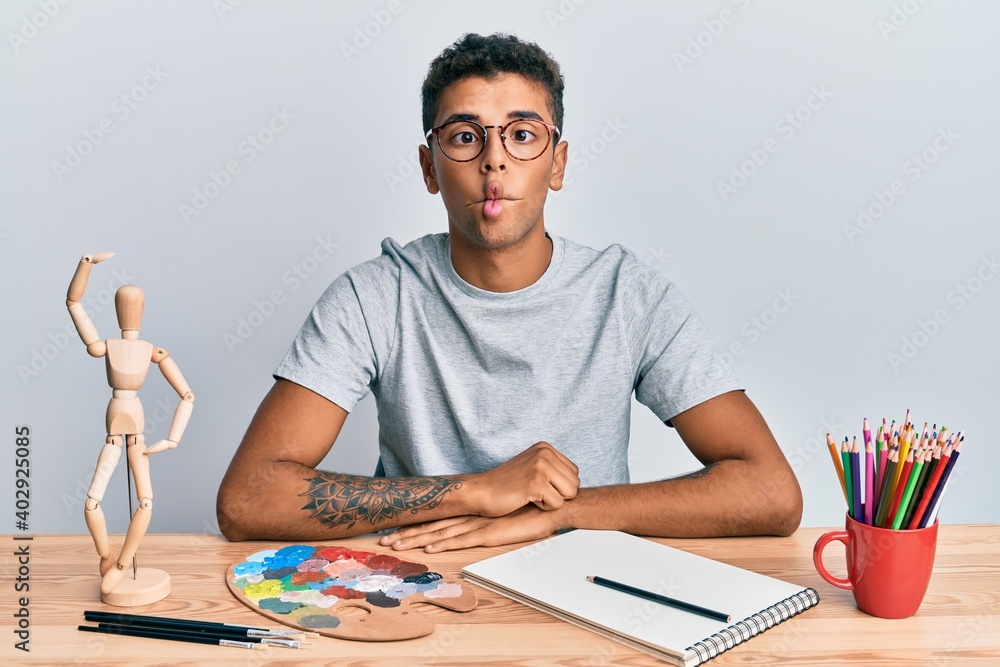 Young handsome african american man painter sitting palette and art manikin making fish face with lips, crazy and comical gesture. funny expression. Young handsome african american man painter sitting palette and art manikin making fish face with lips, crazy and comical gesture. funny expression.