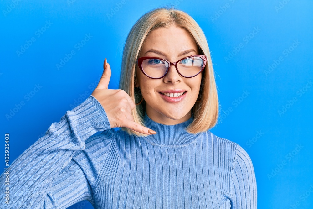 Young blonde woman wearing casual clothes and glasses smiling doing phone gesture with hand and fingers like talking on the telephone. communicating concepts. Young blonde woman wearing casual clothes and glasses smiling doing phone gesture with hand and fingers like talking on the telephone. communicating concepts.